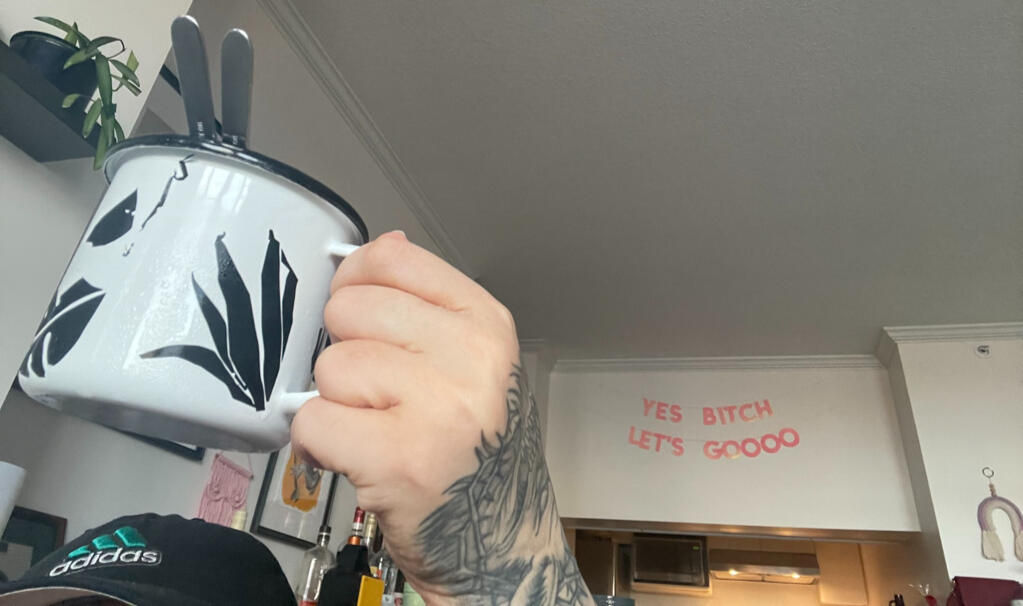 Kristi smilling and sitting sideways in a chair at a cool, cozy coffee shop, underneath a wall mural. Photo by Sasha Stone, July 2024.