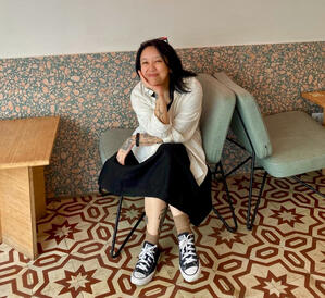 Kristi sits in a comfy chair, with their legs crossed, elbow propped on their knee, resting their chin in their hand and smiling at the camera. They're wearing a black dress, an open white shirt with the sleeves rolled up, and a pair of low-rise Converse.